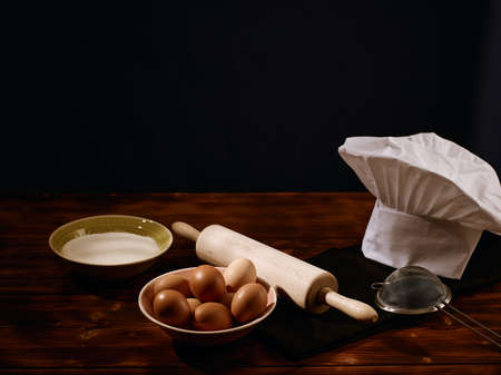 Image of a rolling pin, a sieve, a bowl of flour, eggs in a bowl and chefs hat with copyspace