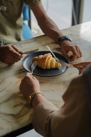 Croissant in a black plate as a man and a woman wanting to cut it with knife and fork.の写真素材