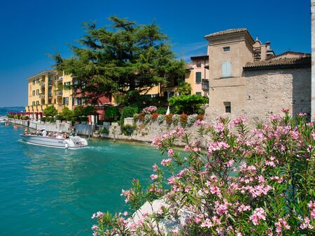 Motorboat just passing under the bridge that leads to Scaligero Castle in the village of Sirmione on the shore of Lake Garda with tourists on board during a summer morning.のeditorial素材