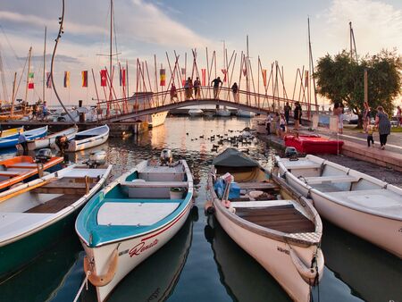 Small fishing boats anchored in the marina of Bardolino and visitors observe a group of ducks from the marina bridge during sunsetのeditorial素材