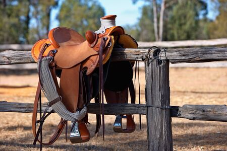 A western style saddle siting upon a country fence beside a riding arena.の写真素材