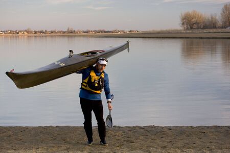 Maturemale paddler in a life jacket is carrying a long racing kayak and a wing paddle on a lake...