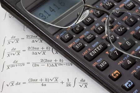 Scientific calculator with pi number on a display and reading glasses against math text book with...