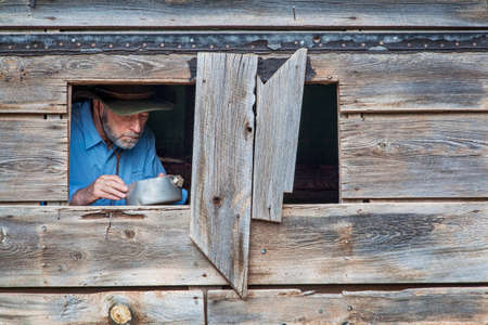 cowboy cooking or eating in an old shack, a face visible through window