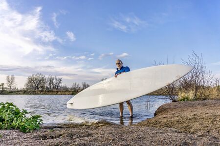 senior male paddler is launching his paddleboard on a lake, early spring in Coloradoの写真素材