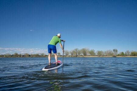 Senior male exercising on stand up paddling (SUP) board.. Early spring on calm lake in Fort Collins, Colorado..の写真素材