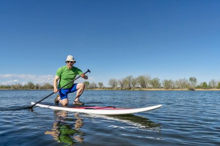 Senior male exercising on stand up paddling (SUP) board. Early spring on a calm lake in Fort Collins, Colorado..の写真素材