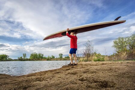 senior male paddler carrying his SUP paddleboard on a lake shore in Colorado, early springの写真素材