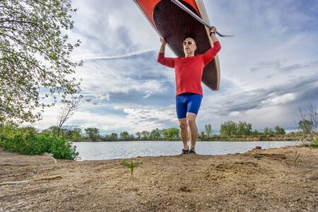 senior male paddler carrying his SUP paddleboard on a lake shore in Colorado, early springの写真素材