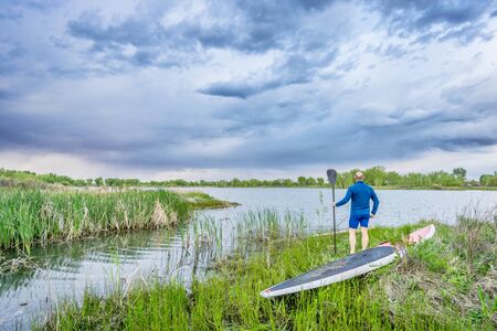 senior male with stand up paddleboard on lake shore watching stormy clouds