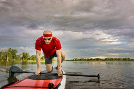 senior male paddler starting his workout on  stand up paddleboard, a local lake in Colorado under cloudy skyの写真素材