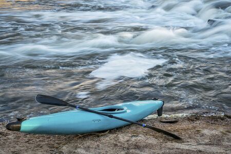 blue plastic white water kayak with a paddle on a river shore with a rapid in backgroundの写真素材