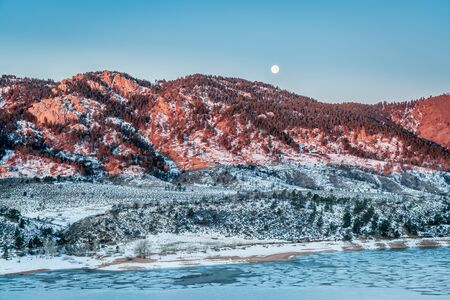 Moon setting over Arhturs Rock lit by sunrise with Horsetooth Reservoir covered by ice, Fort Collins, Coloradoの写真素材