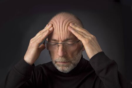tired - 60 years old  man with a beard and glasses massaging his forehead -  a headshot against a black backgroundの写真素材