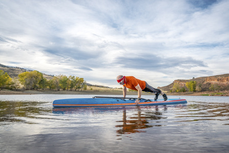 A senior male exercising (pushups and planks) on a stand up paddleboard, a calm mountain lake in fall colorsの写真素材