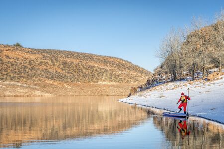 male paddler in drysuit is launching a stand up paddleboard on mountain lake in Colorado, winter sceneryの写真素材