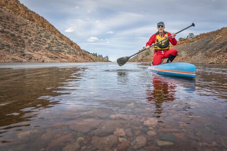 senior male paddler in drysuit  is enjoying stand up paddling on lake in Colorado, winter scenery with some iceの写真素材