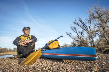 a senior male paddler is taking a rest on a gravelbar during stand up paddling on the South Platte RIver, cold season sceneryの写真素材