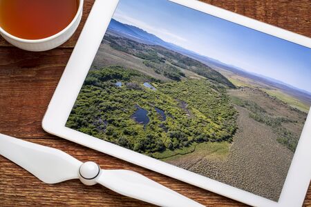 reviewing aerial pictures of a stream at foothills of Medicine Bow Mountains (North Park, Colorado) on a digital tablet with a cup of teaの写真素材