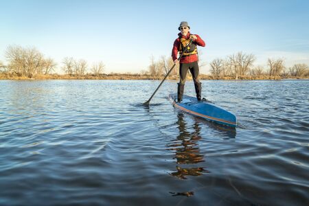 senior male paddling a paddleboard on a lake in Colorado, winter or early spring sceneryの写真素材