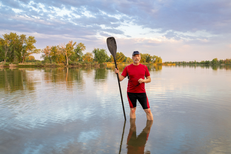 Senior male paddler with a long stand up paddle standing in a calm lake, summer sceneryの写真素材