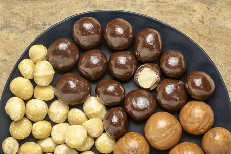 macadamia nuts in shells, shelled and dipped in dark chocolate on a black plateの写真素材
