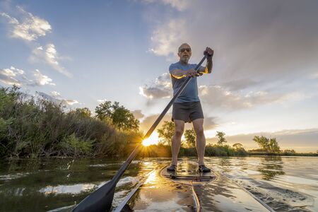 senior male paddler is paddling stand up paddleboard at sunset on a lake in Coloradoの写真素材