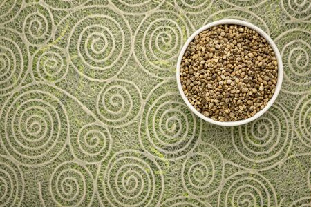 dry hemp seeds in a small ceramic bowl, top view against lace paper with spiral patternの写真素材