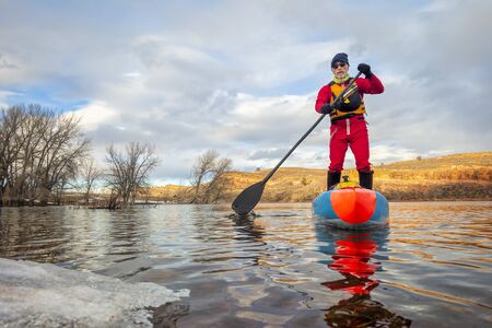 senior paddler in a dry suit and life jacket is training on stand up paddleboard on a mountain lake in winter conditions - Horsetooth Reservoir in northern Colorado, POV from an action cameraの写真素材