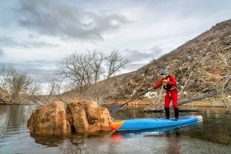 senior male paddler in a dry suit and life jacket is navigating his paddleboard between rocks, trees...