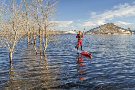 senior male paddler in a drysuit and life jacket is paddling a long unlimited stand up paddleboard in winter conditions on a lake in Colorado - Horsetooth Reservoir, fitness and training conceptの写真素材