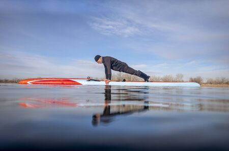 senior male paddler wearing wetsuit is doing push ups on a stand up paddleboard, calm lake in Colorado, early spring, training and fitness conceptの写真素材