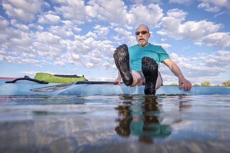 senior male paddler wearing five finger water shoes is sitting on his stand up paddleboard - low...