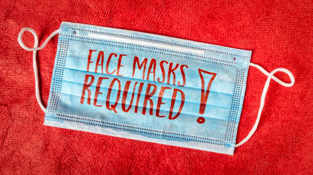 face masks required - text on a disposable mask against red textured paper, business sign during the coronavirus covid-19 pandemic and social distancingの写真素材