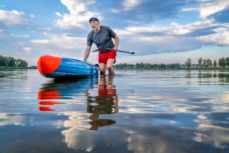 senior male stand up paddler is launching his paddleboard  on a lake for morning workout in northern Colorado, solo paddling as a form of social distancingの写真素材