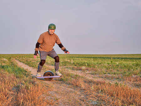 sunrise ride on a prairie, mature senior male is riding one-wheeled electric skateboard on a dirt roadの写真素材