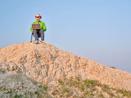 Senior man in cowboy hat is sitting on a folding chair and working on laptop in the middle of nowhere, early morning in the badlands of Pawnee National Grassland in Coloradoの写真素材