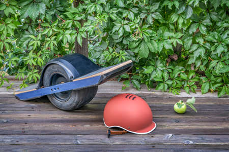 scratched and dirty one-wheeled electric skateboard with a helmet on a wooden deck with green vineの写真素材