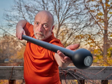 senior man is exercising with a steel mace in his backyard, selective focus with fall sceneryの写真素材