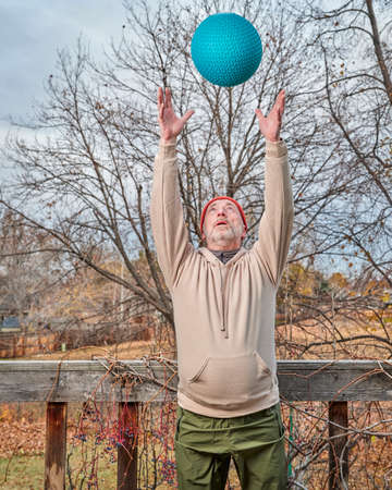 senior man (in late 60s) is exercising with a heavy slam ball in his backyard, chilly afternoon in...