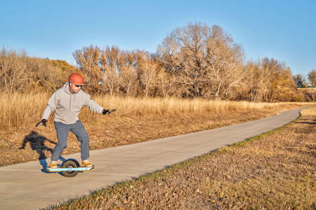 senior man is riding one-wheeled electric skateboard on a paved bike trail in fall scenery in...