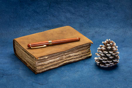 antique leather-bound journal with decked edge handmade paper pages with a stylish pen and a decorative pine cone against blue handmade paper, journaling or winter holiday greetings conceptの写真素材