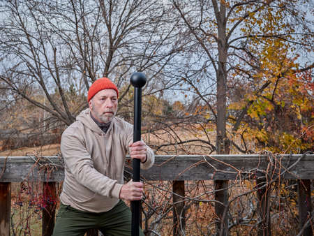 senior man (in late 60s) is exercising with a steel mace in his backyard, fall scenery