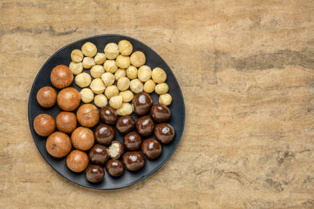 macadamia nuts in shells, shelled and dipped in dark chocolate on a black plate against buckskin textured paper background with a copy spaceの写真素材