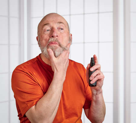 bald and bearded senior man is making funny faces when applying grooming conditioner oil to his beard, self care for men conceptの写真素材