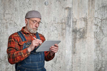confident senior man wearing glasses, a flannel shirt and overalls is making notes or sketches on a...