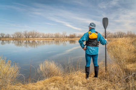 senior male stand up paddler wearing life jacket on a shore of calm lake in early spring scenery in...