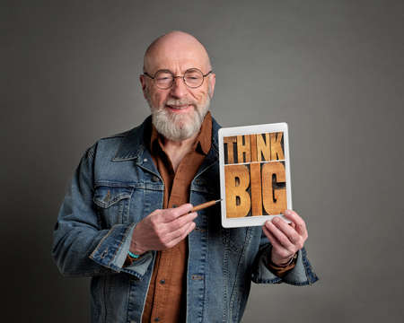 smiling senior man is showing a digital tablet with think big sign in retro letterpress wood type, optimism and attitude conceptの写真素材