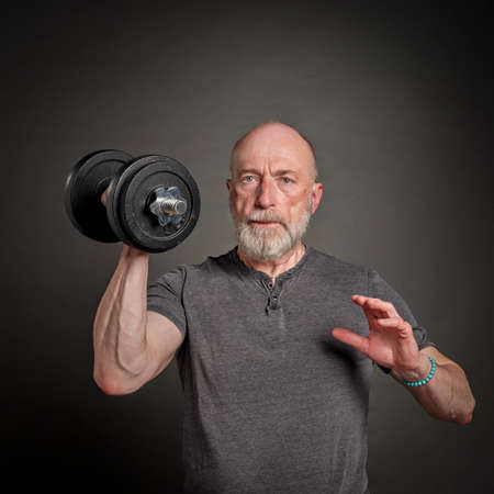 casual portrait of old bearded  man (in late 60s) exercising with heavy dumbbell, active senior and...
