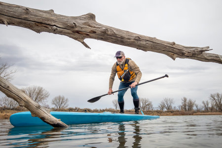 senior male paddling a stand up paddleboard on a lake in early spring, frog perspective from an...
